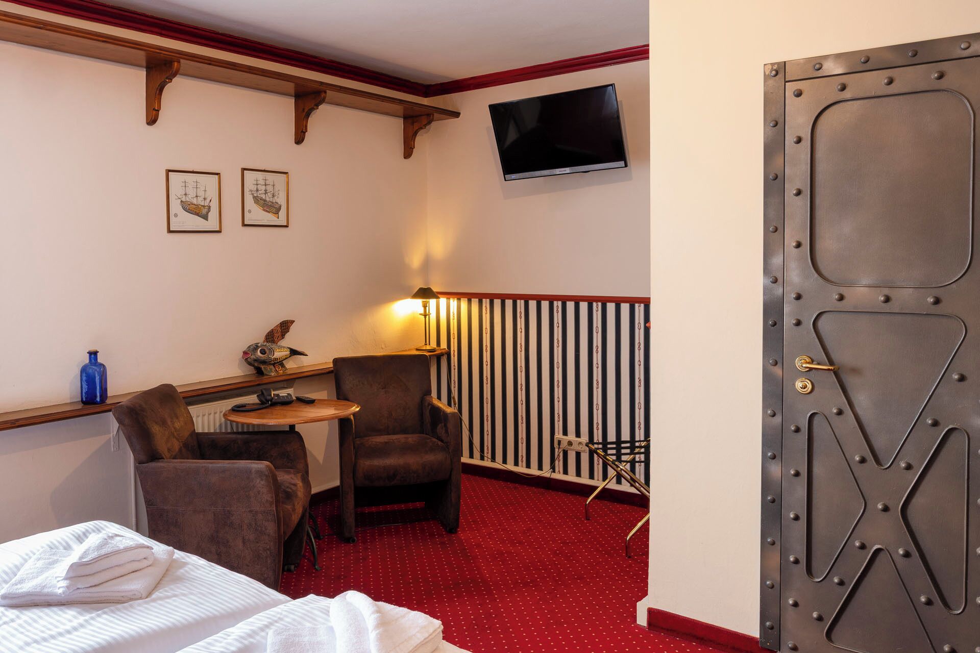 A hotel room with a bed, a desk with a lamp, two chairs, a television on the wall, and a large metal door.