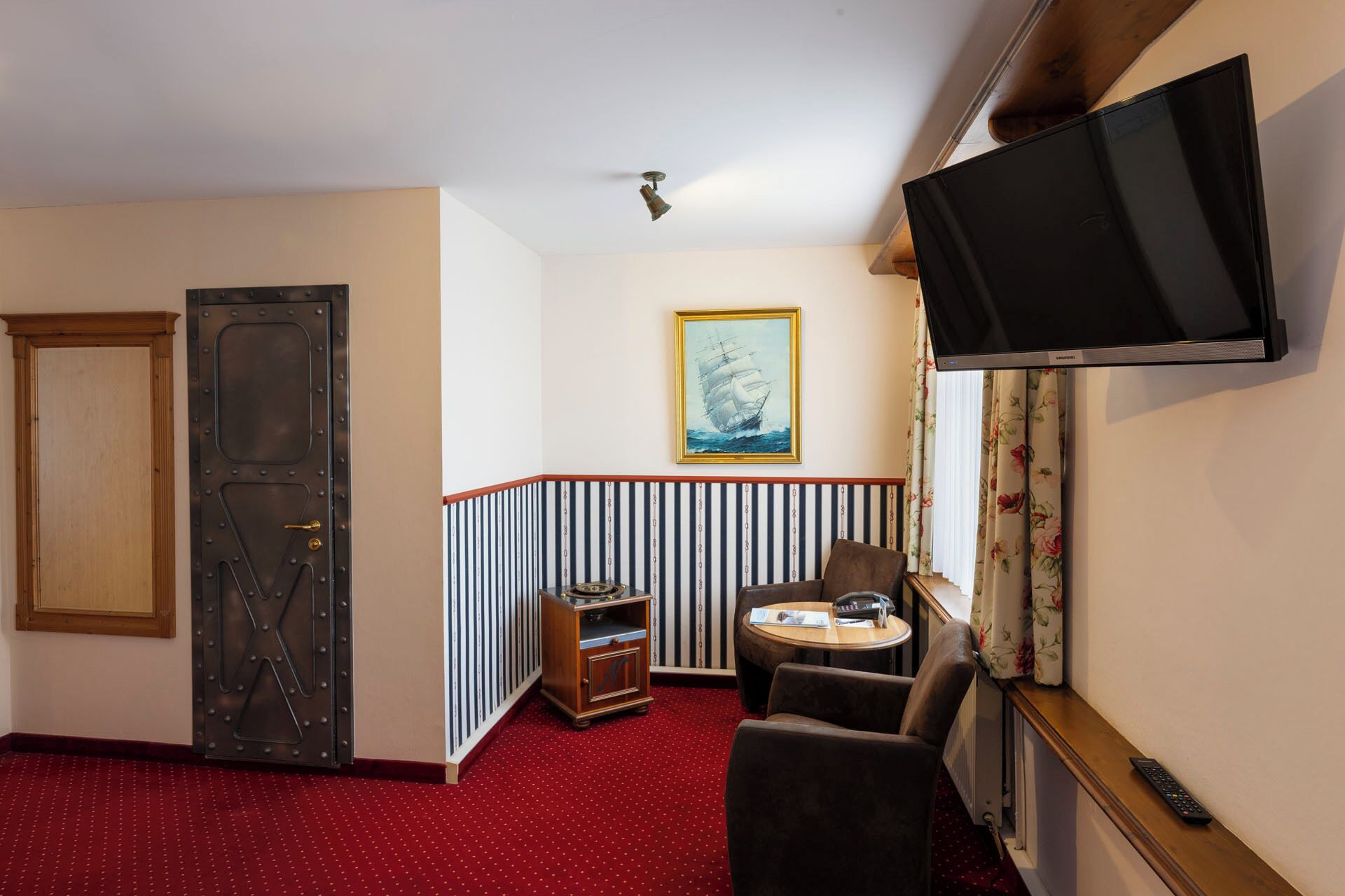 A hotel room with a red carpet, a wooden door, a television, a framed picture, a small table, and a chair.