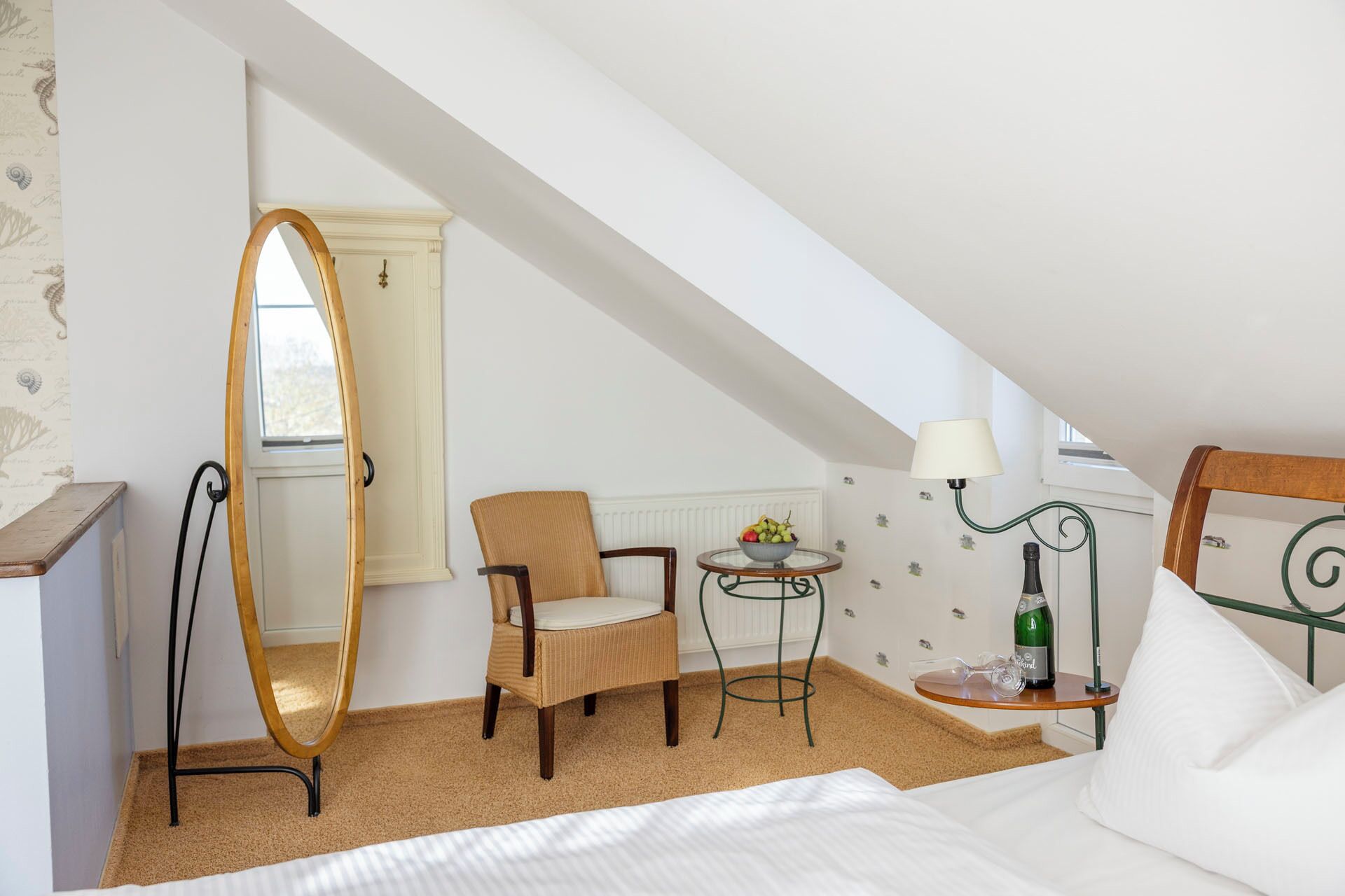 Una habitación pequeña y acogedora con una cama, una silla, una mesa redonda con lámpara y un espejo.