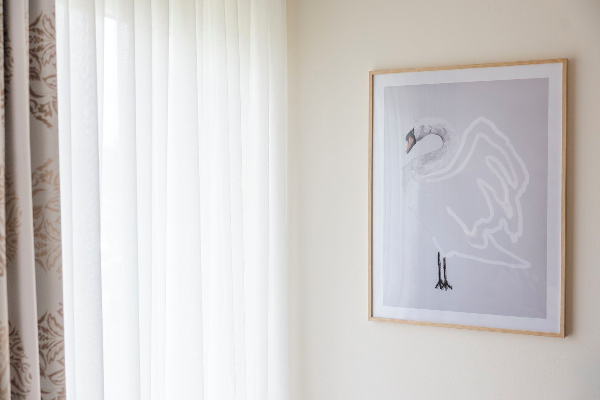 A framed abstract artwork on a wall, a window with sheer curtains, and a plain wall.