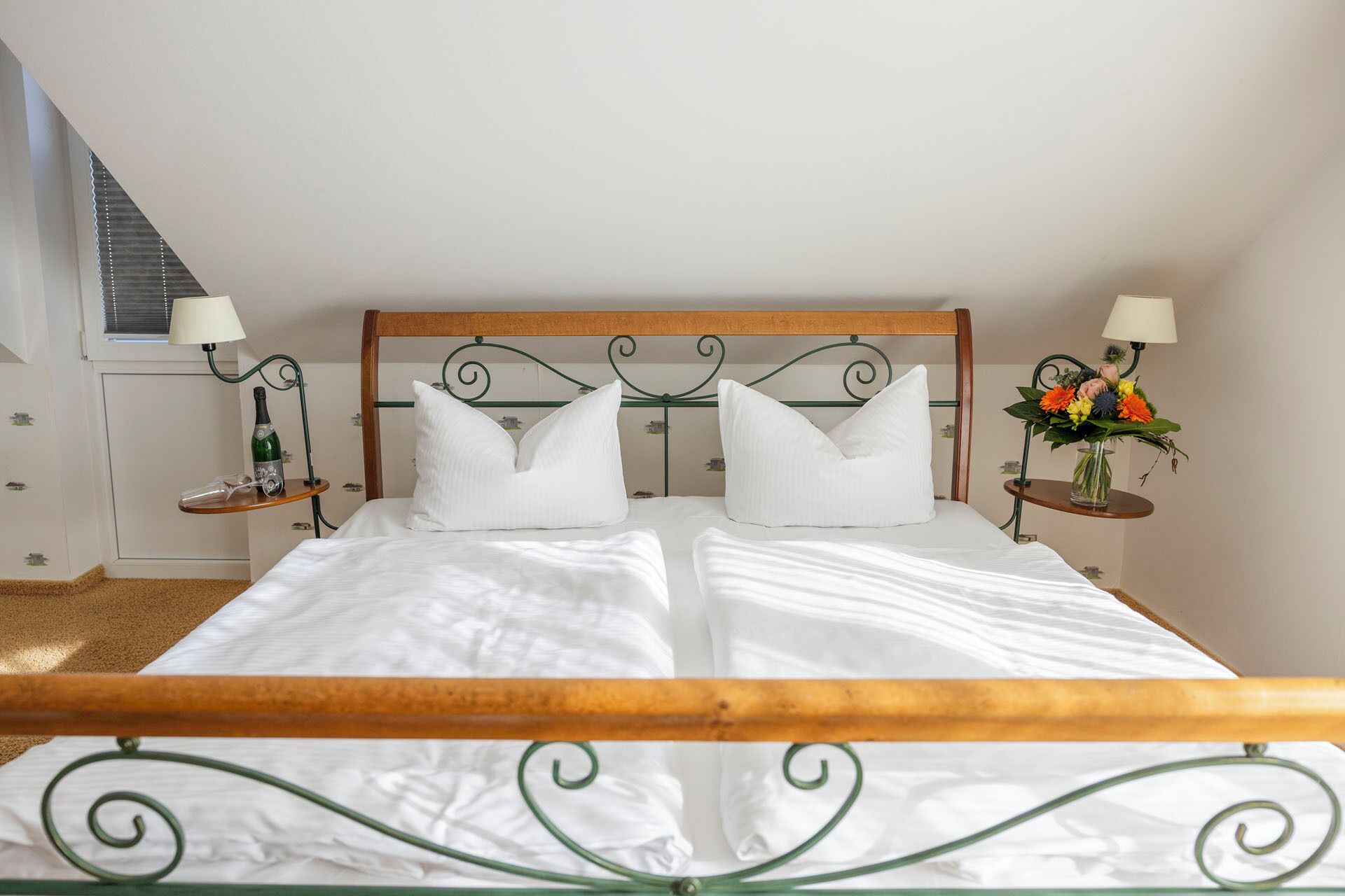 Een net opgemaakt bed met een houten hoofdbord en metalen frame, met aan beide zijden nachtkastjes, lampen en een bloevas.