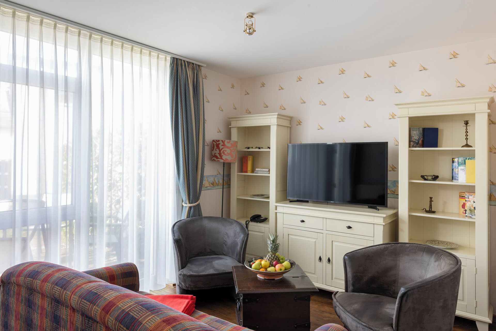 Een woonkamer met een bank, twee fauteuils, een salontafel met fruit, een televisie en een boekenkast.