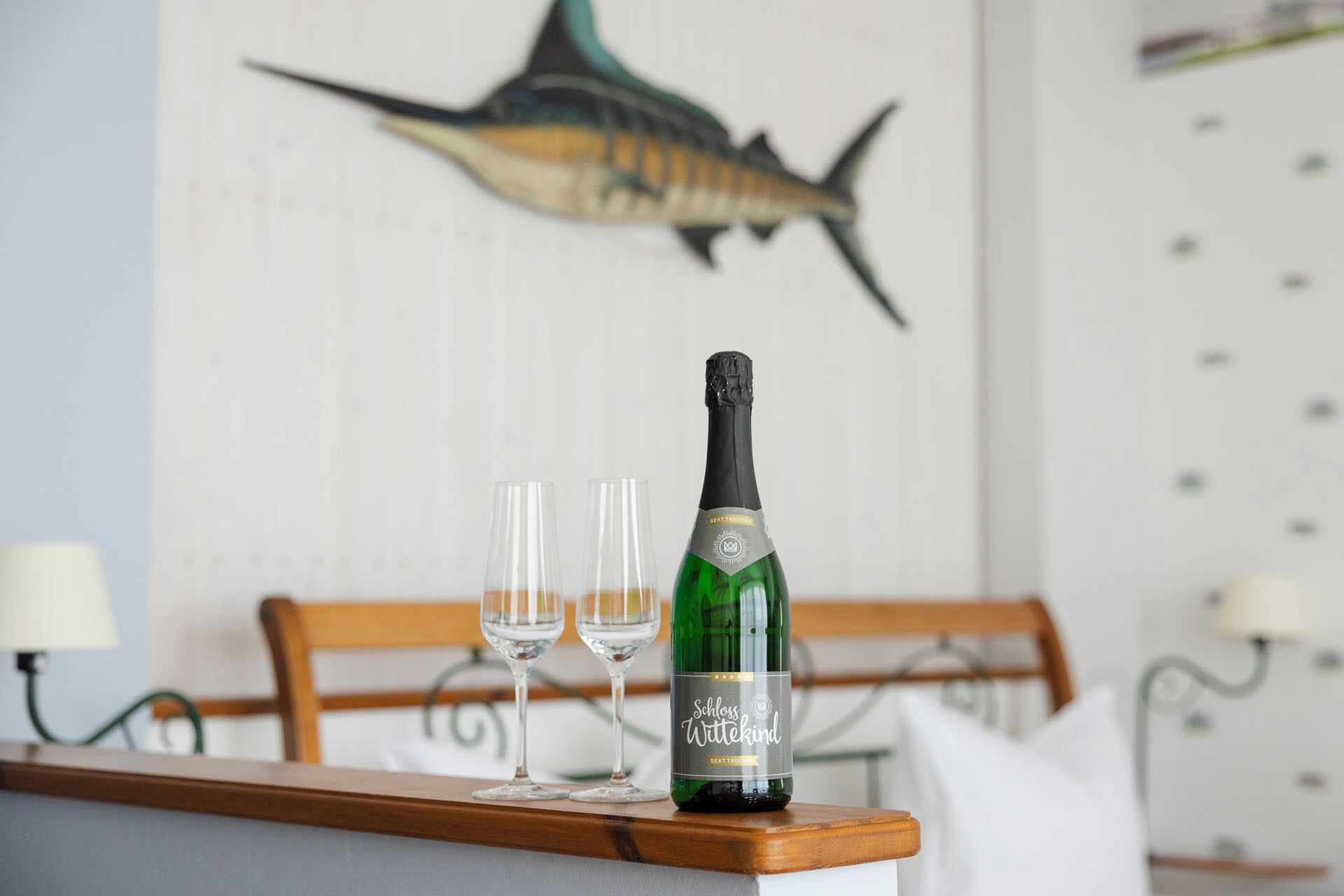 Uma garrafa de vinho Schloss Wuteking e dois copos vazios sobre uma mesa de madeira num quarto com uma cama e uma decoração de peixe na parede.