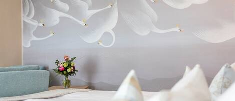 A bedroom with a large wall mural of swans, a bed with white bedding, a vase with flowers, and a blue armchair.