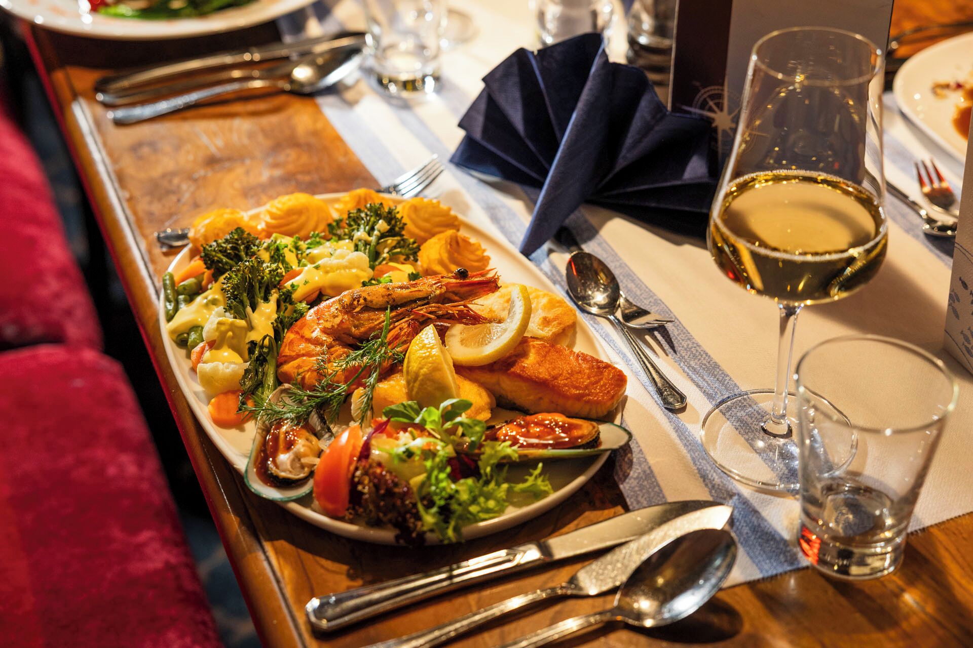 A dining table with a variety of seafood and vegetables, a glass of white wine, and dining utensils.