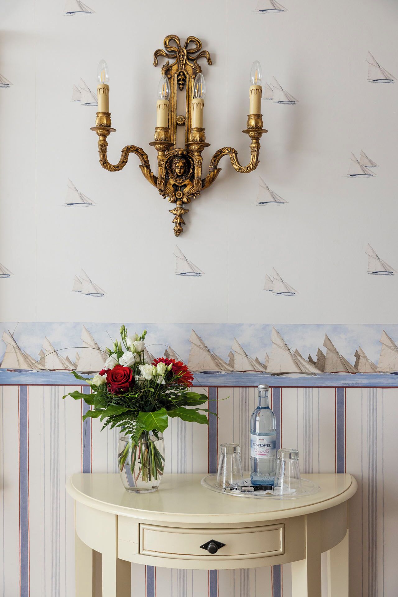 Un candelabro dorado sujeto a la pared con tres velas, un florero con flores y una botella sobre una mesa blanca, con un fondo de papel tapiz a rayas.