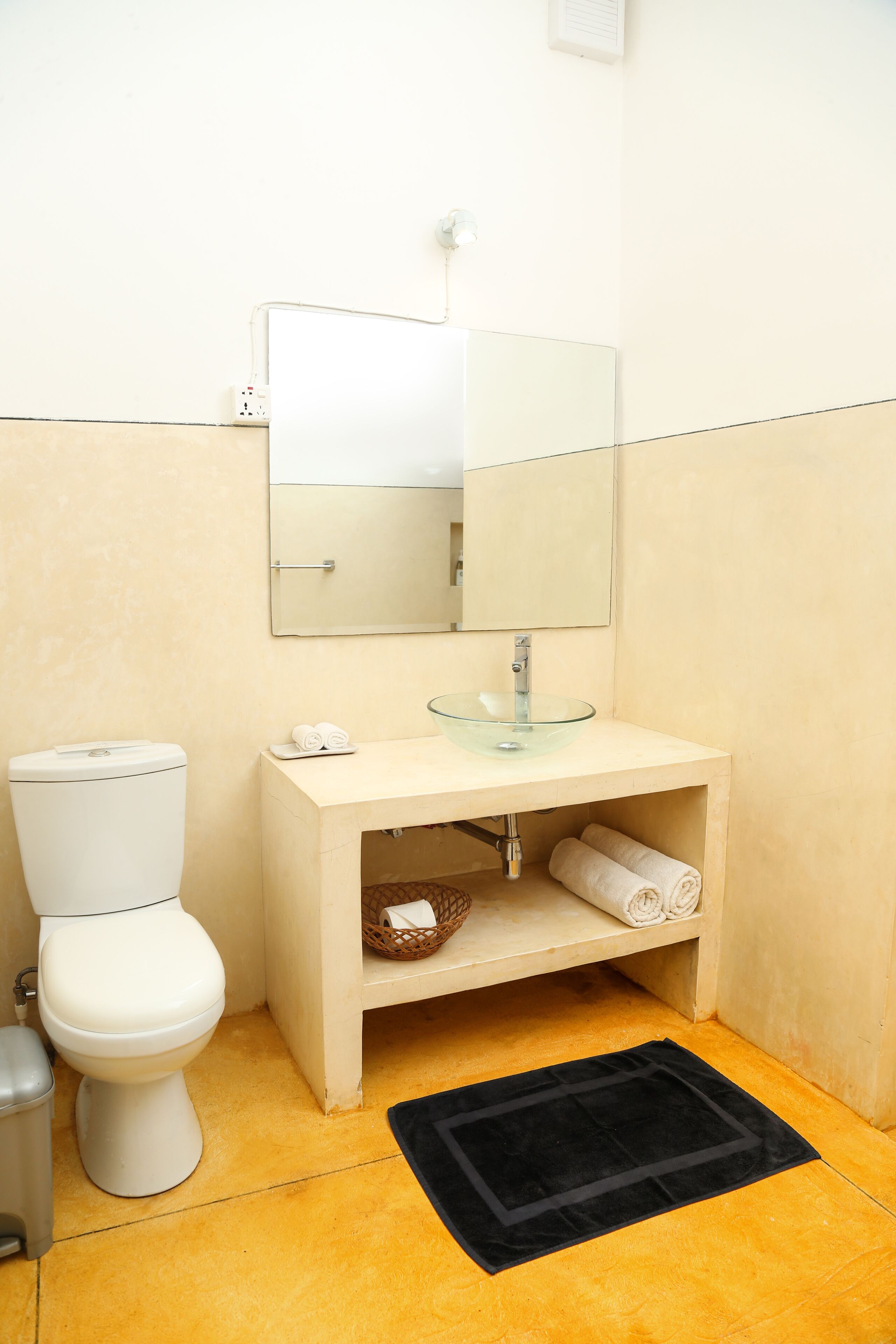 A bathroom with a toilet, a glass sink, a mirror, and a black mat on the floor.