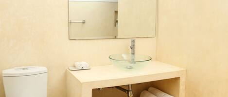 A bathroom with a toilet, a glass sink, a mirror, and a black mat on the floor.