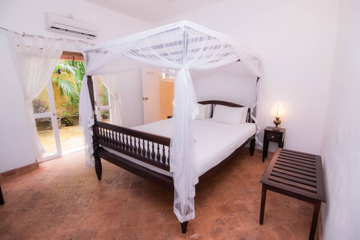 Un dormitorio con cama con dosel, una mesita de noche con lámpara y vista al jardín a través de la puerta.