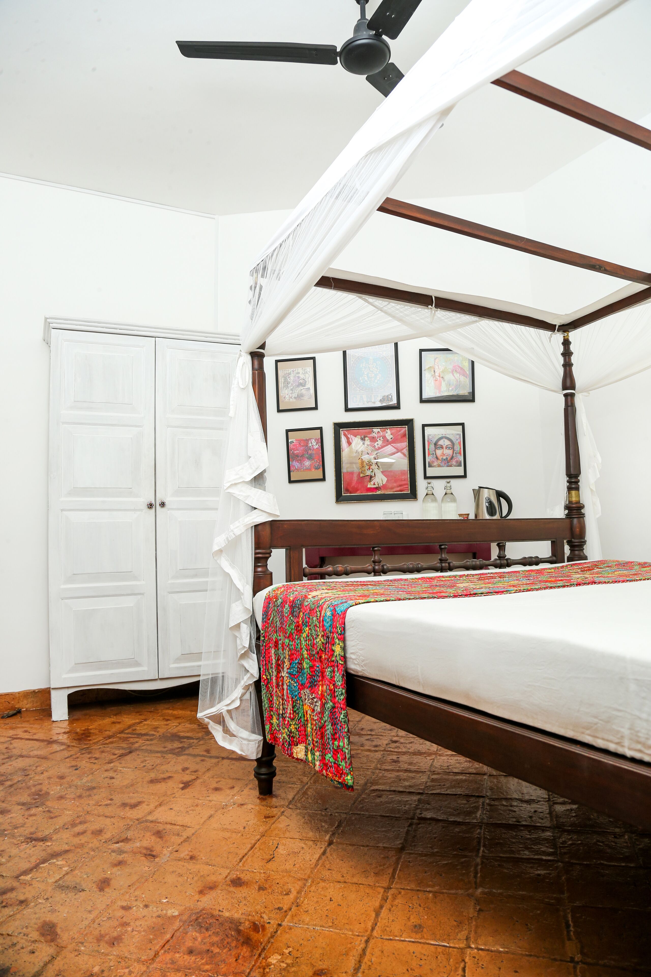 Una cama con dosel y cuatro columnas con una colcha blanca, un cabecero de madera y un colorido plaid. Hay cuadros enmarcados en la pared, un armario blanco y un ventilador de techo.