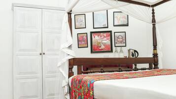 Una cama con dosel y cuatro columnas con una colcha blanca, un cabecero de madera y un colorido plaid. Hay cuadros enmarcados en la pared, un armario blanco y un ventilador de techo.