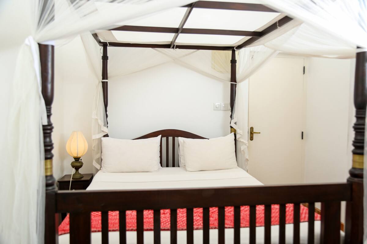 Una cama con dosel y cuatro columnas, con ropa de cama blanca y una colcha a rayas rojas y blancas.