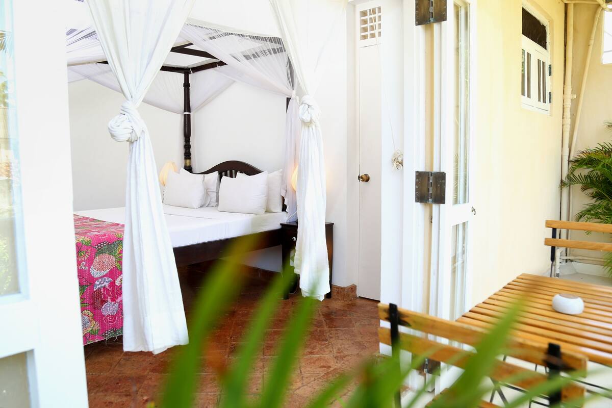 Habitación con cama bajo un dosel blanco, un banco de madera y una puerta con ventanal.