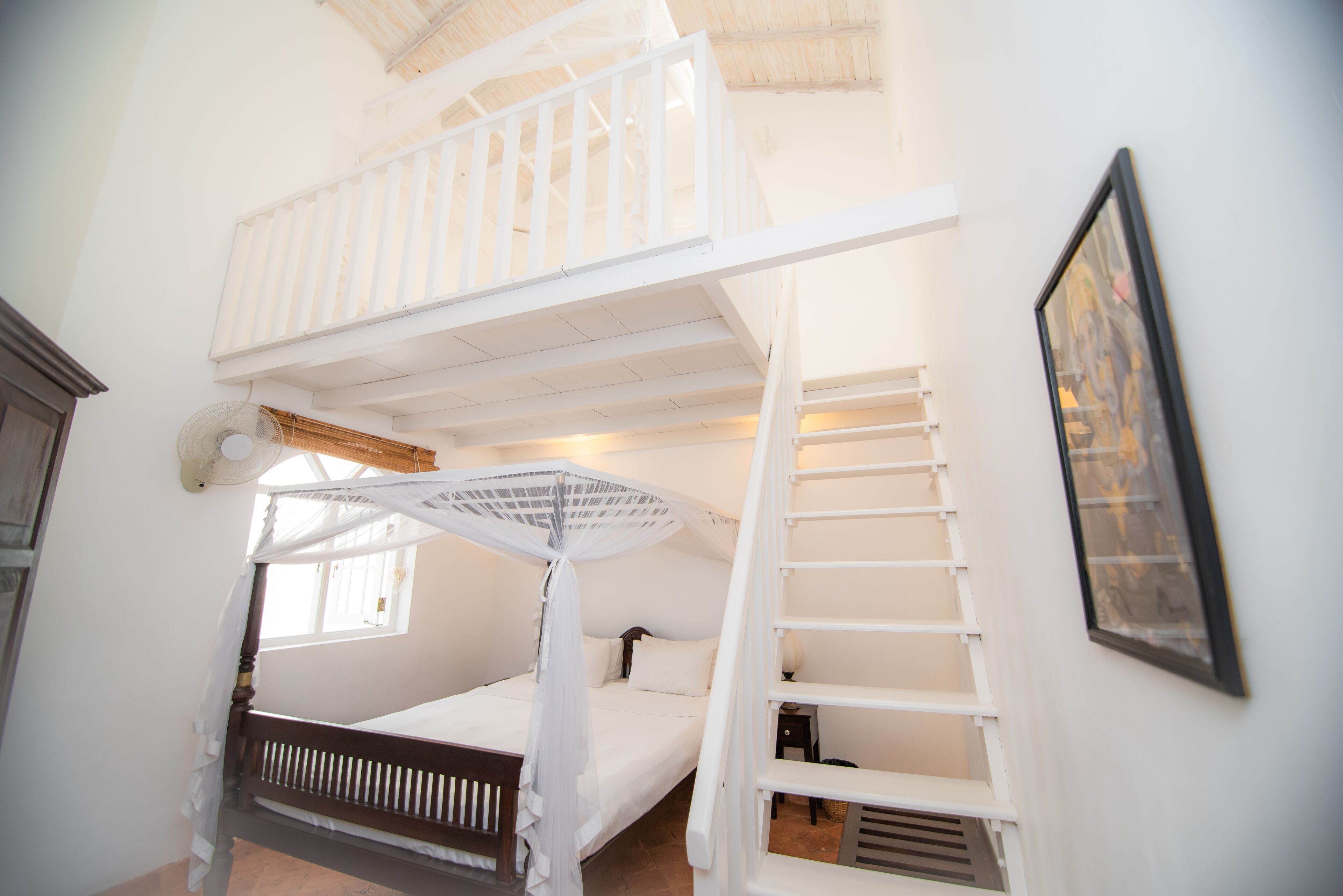 Een kamer met een stapelbed, een ladder, een raam en een ingelijst schilderij aan de muur.