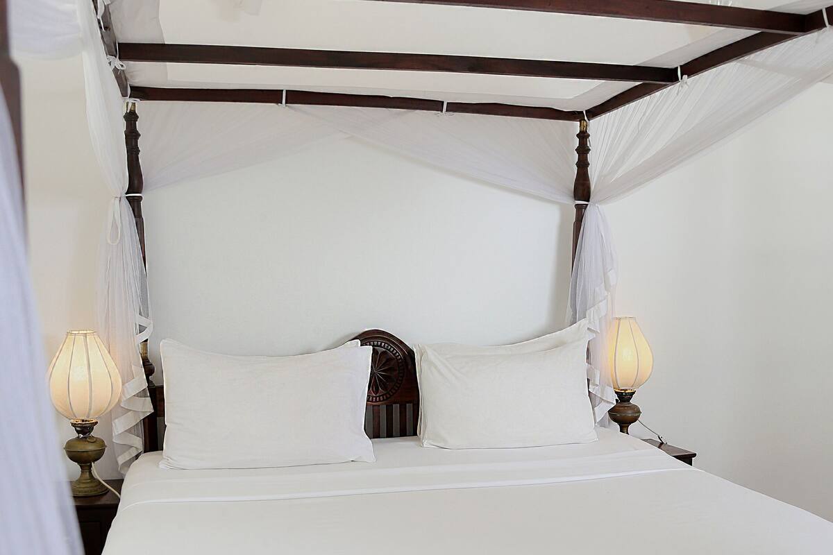 Una cama con dosel y cuatro columnas, con sábanas blancas, una colcha de flores y dos lámparas de noche.