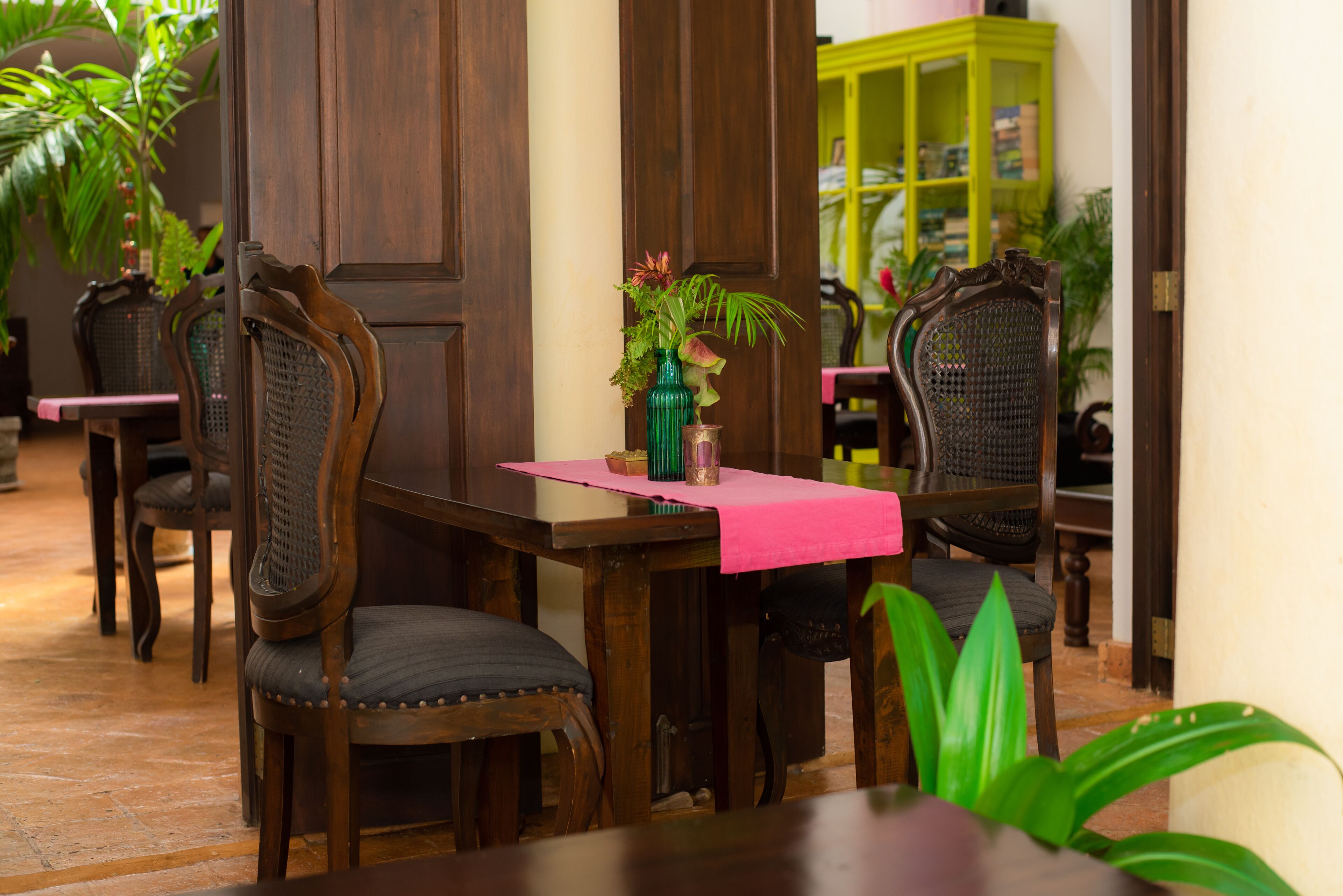 A dining area with wooden tables and chairs, a green vase with flowers, and a yellow cabinet in the background.