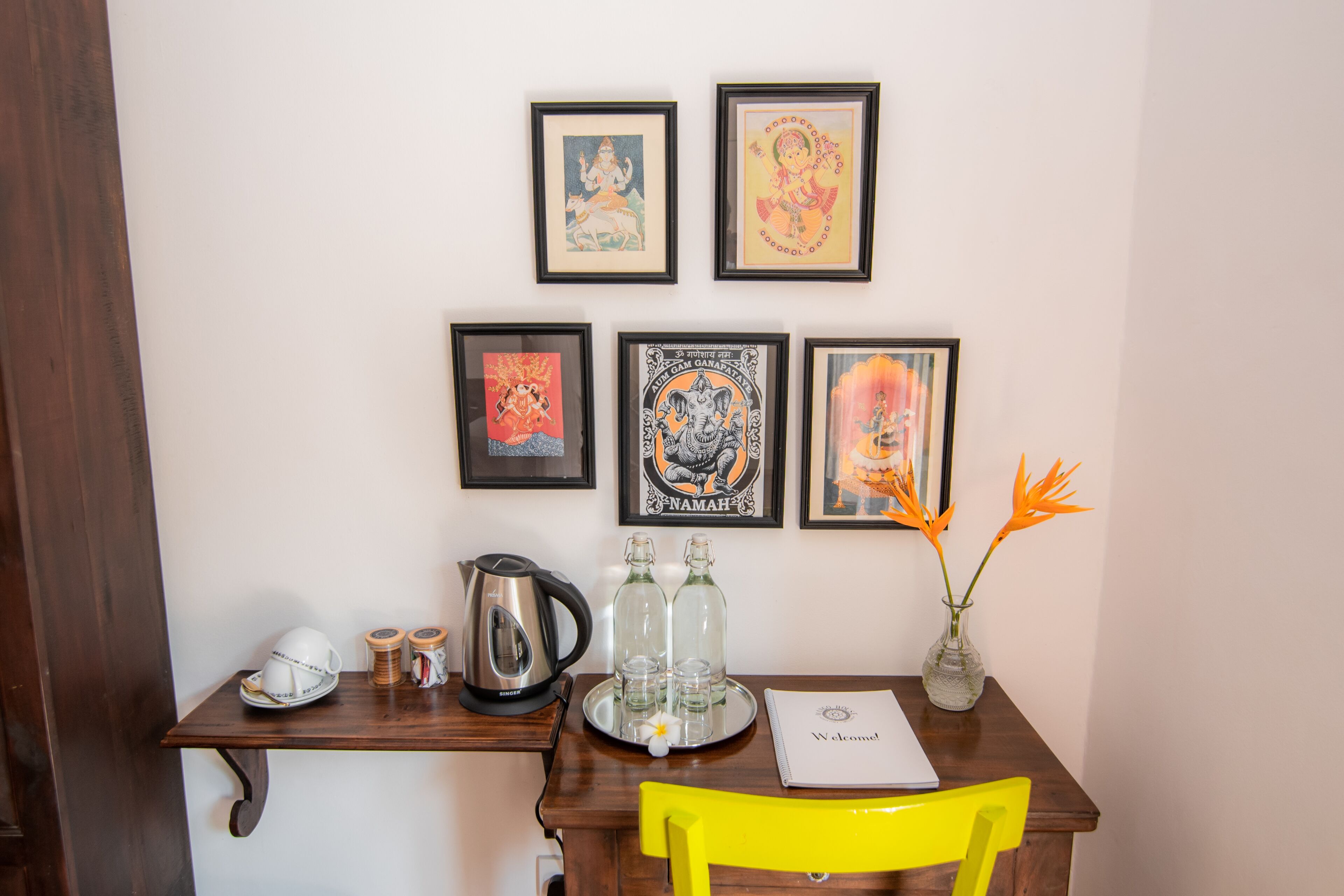 Una habitación con un escritorio de madera, una silla amarilla, obras de arte enmarcadas en la pared, una bandeja con vasos y una jarrera, y un cartel de bienvenida.