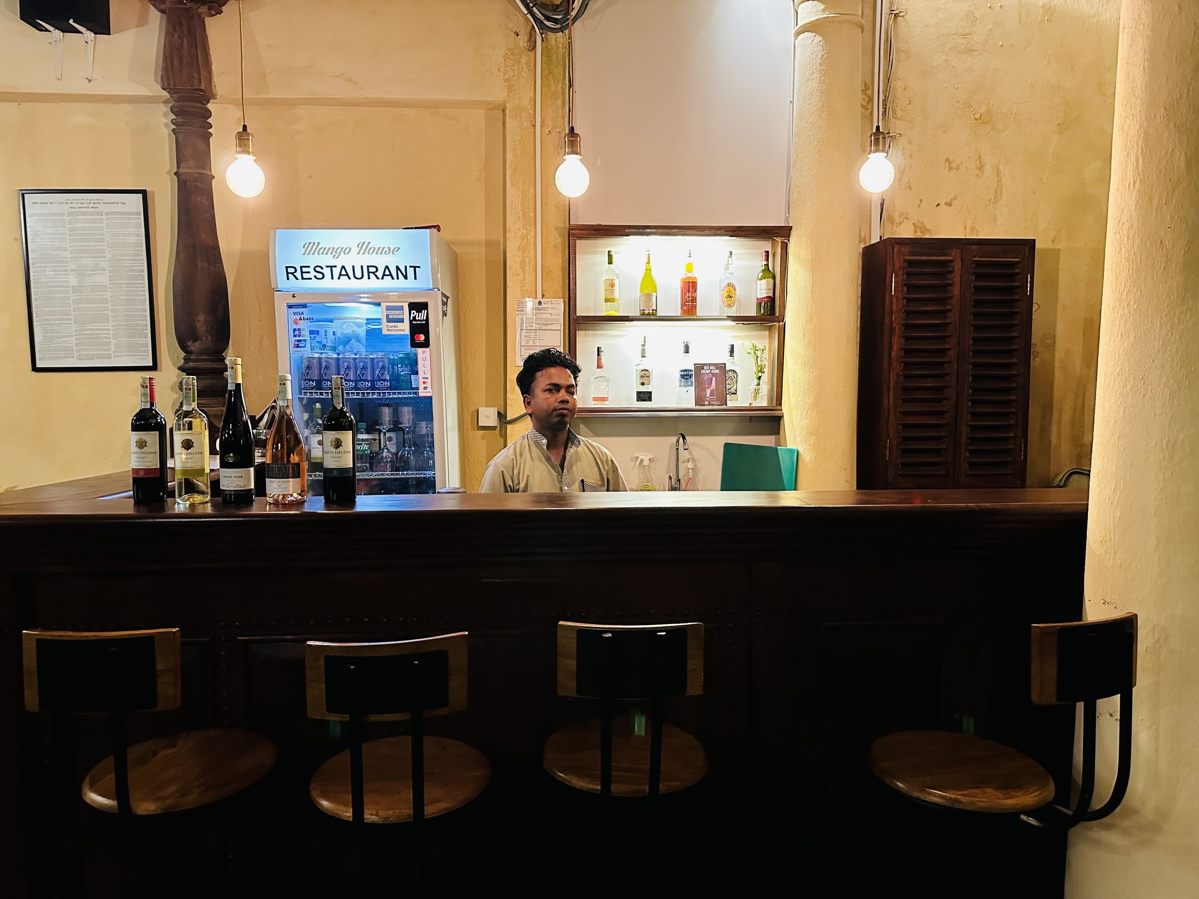 A bar area with a counter displaying various bottles, a sign for 'Mango House Restaurant', and a shelf with bottles in the background.