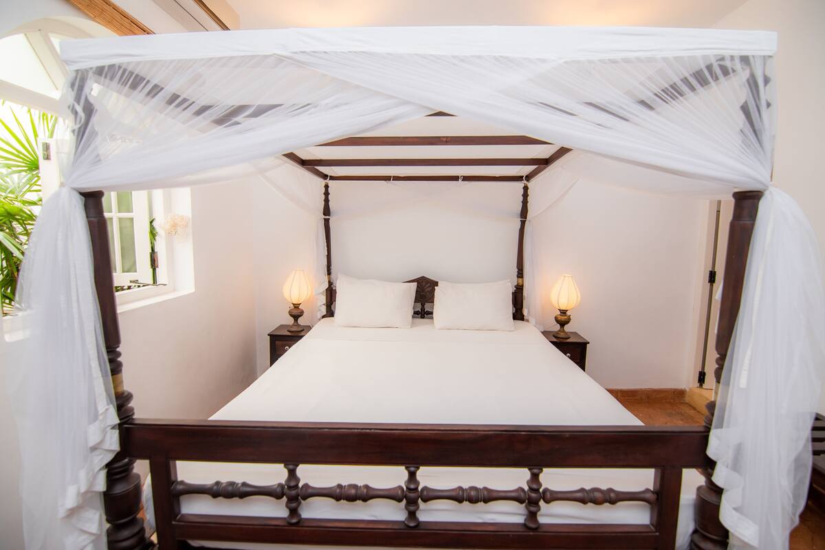 Una cama con dosel adornada con sábanas blancas, dos lámparas de noche y una ventana con vistas a la vegetación.