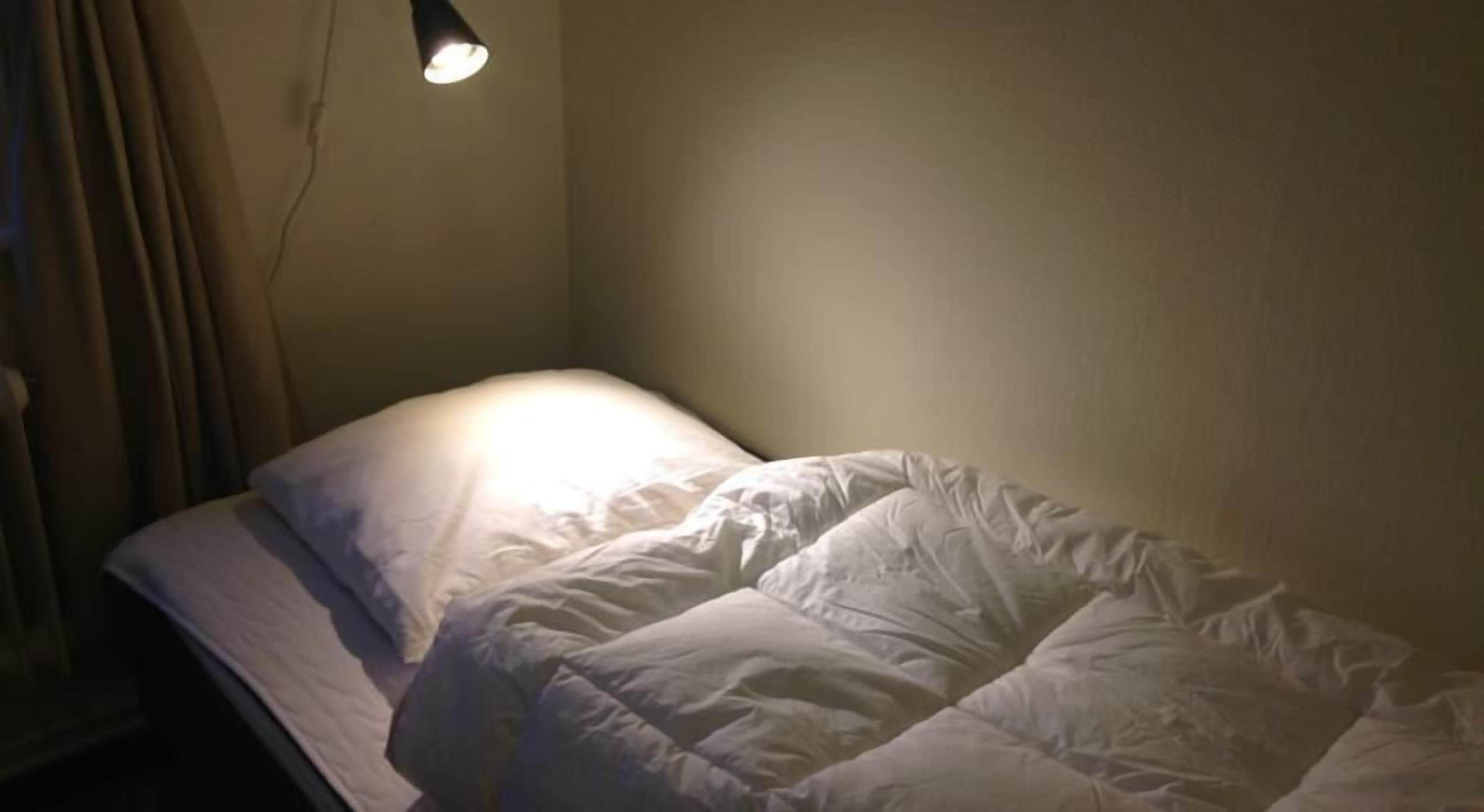 Un lit simple avec une couette blanche et un seul oreiller, une lampe de chevet et un rideau tiré partiellement sur le côté.