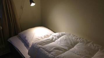Un lit simple avec une couette blanche et un seul oreiller, une lampe de chevet et un rideau tiré partiellement sur le côté.