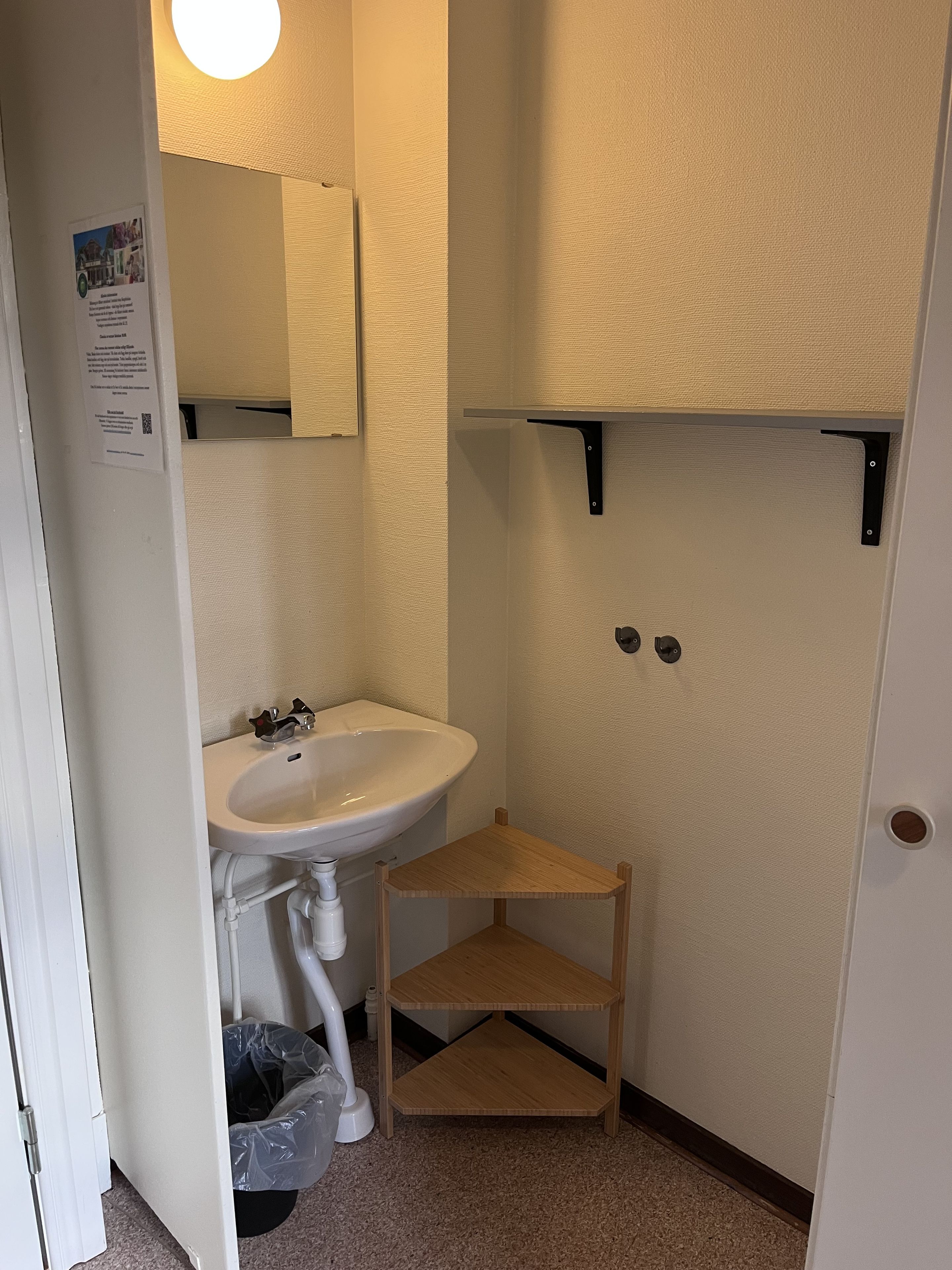 A small bathroom with a white sink, a mirror, a wooden shelf, and a trash bin.