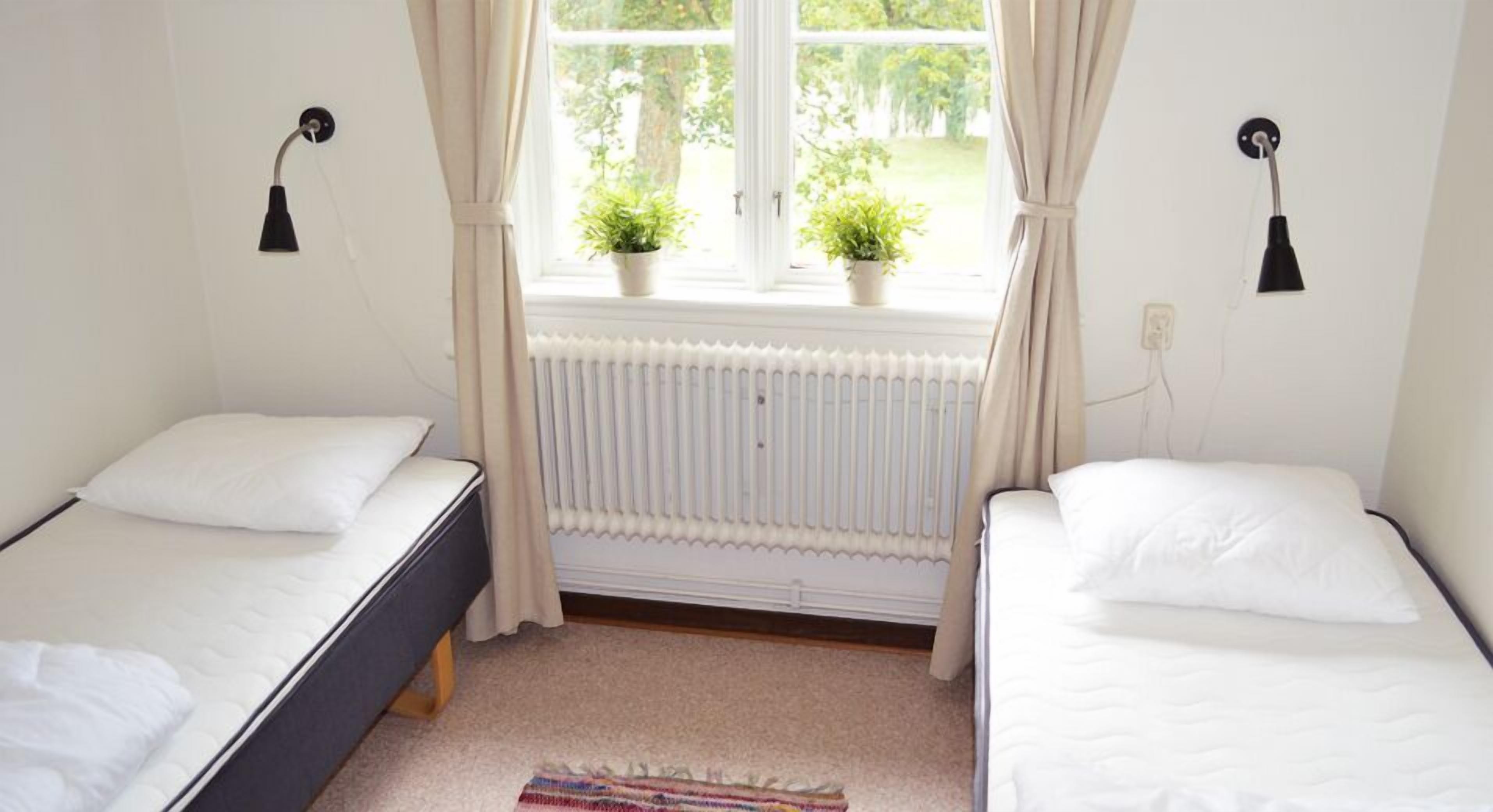 Un dormitorio con dos camas individuales, una ventana con cortinas, un radiador y dos lámparas fijadas a la pared.