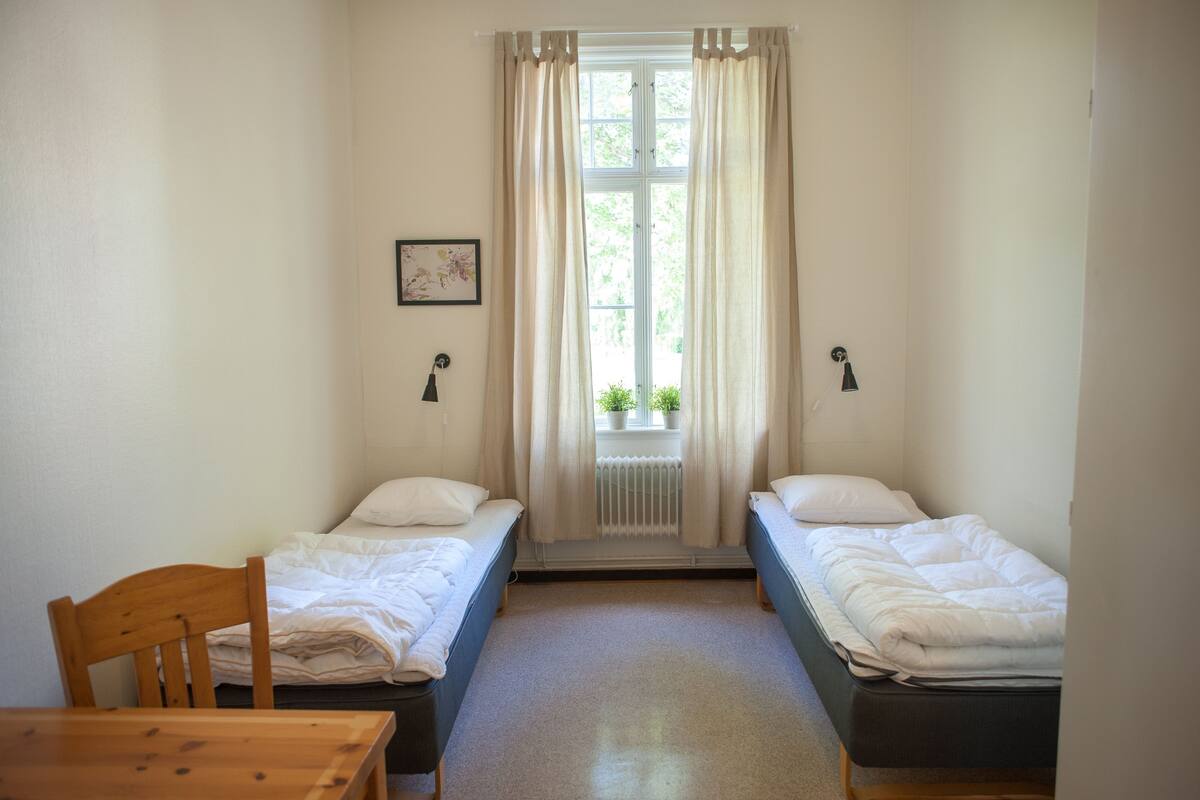 A small, clean room with two single beds, a wooden chair, a table, and a window with curtains.