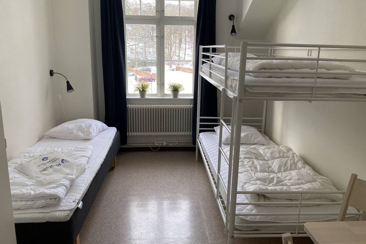 A dormitory room with bunk beds, a window with curtains, a radiator, and a small table.