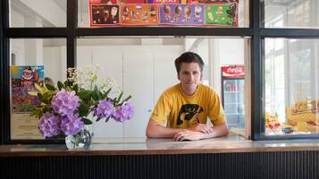 A person seated at a counter with a vase of flowers, a menu board above, and a Coca-Cola refrigerator in the background.