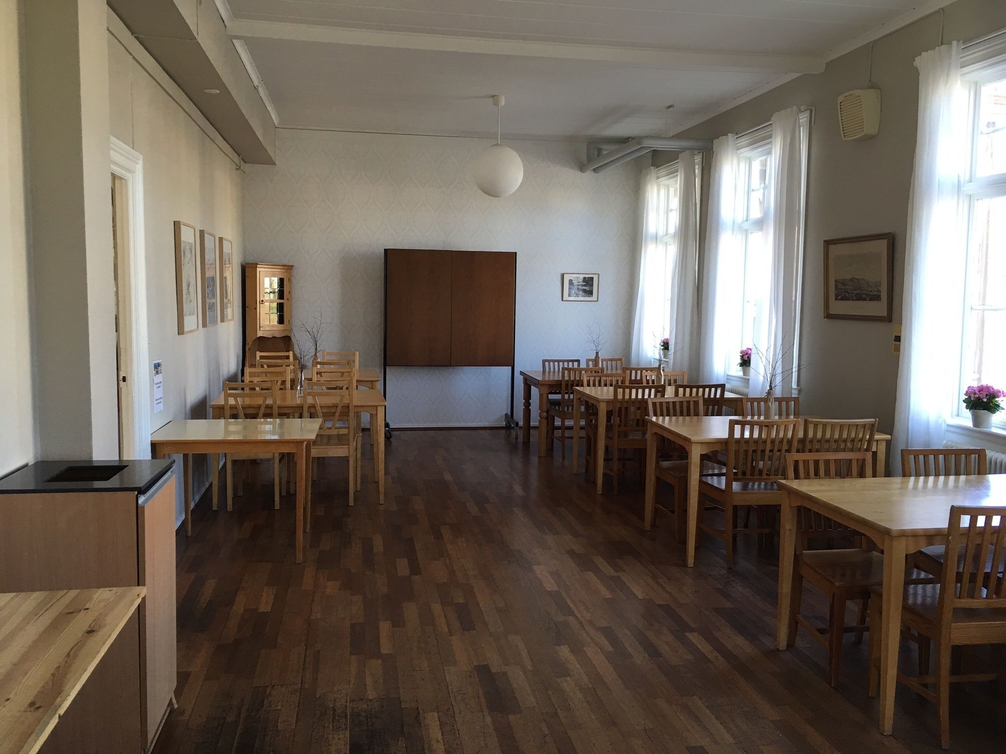 A spacious room with wooden flooring, multiple tables and chairs, a cabinet, and a large window with curtains.