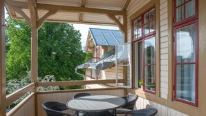 A covered outdoor area with a round table and wicker chairs, a wooden deck, and a view of a house with solar panels and red-framed windows. - Ronneby Brunnspark Vandrarhem och B&B - Hostel (Ronneby)