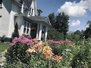 A two-story house with a porch, surrounded by a garden with various flowers and plants. - B&B Hillhouse (Lac-Brome)