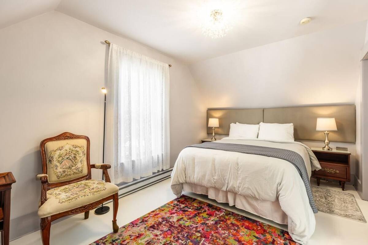 A bedroom with a large bed, two bedside tables with lamps, a chair, a colorful rug, and a window with sheer curtains.