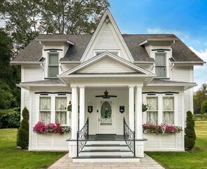 Een wit, twee verdiepingen tellend huis met een centrale ingang, een voortuin en bloembakken aan weerszijden.