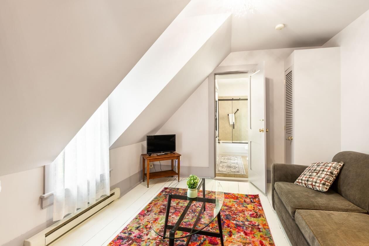 A loft-style living room with a sloped ceiling, a television on a wooden stand, a glass coffee table, a colorful rug, a sofa with a plaid pillow, and a door leading to a bathroom.