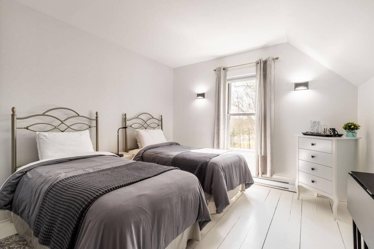 A bedroom with two beds, a white dresser, a window with curtains, and wall-mounted lights.