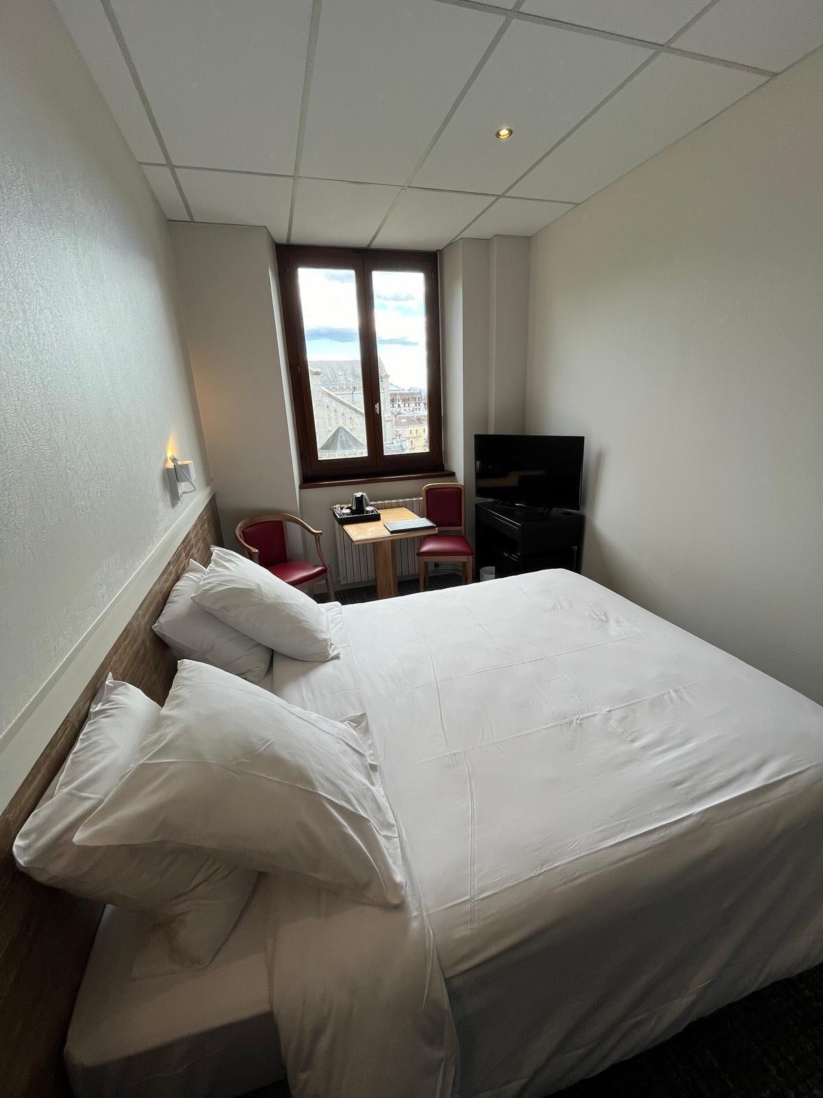 A hotel room with a bed, a desk with a chair, a television, and a window with a city view.