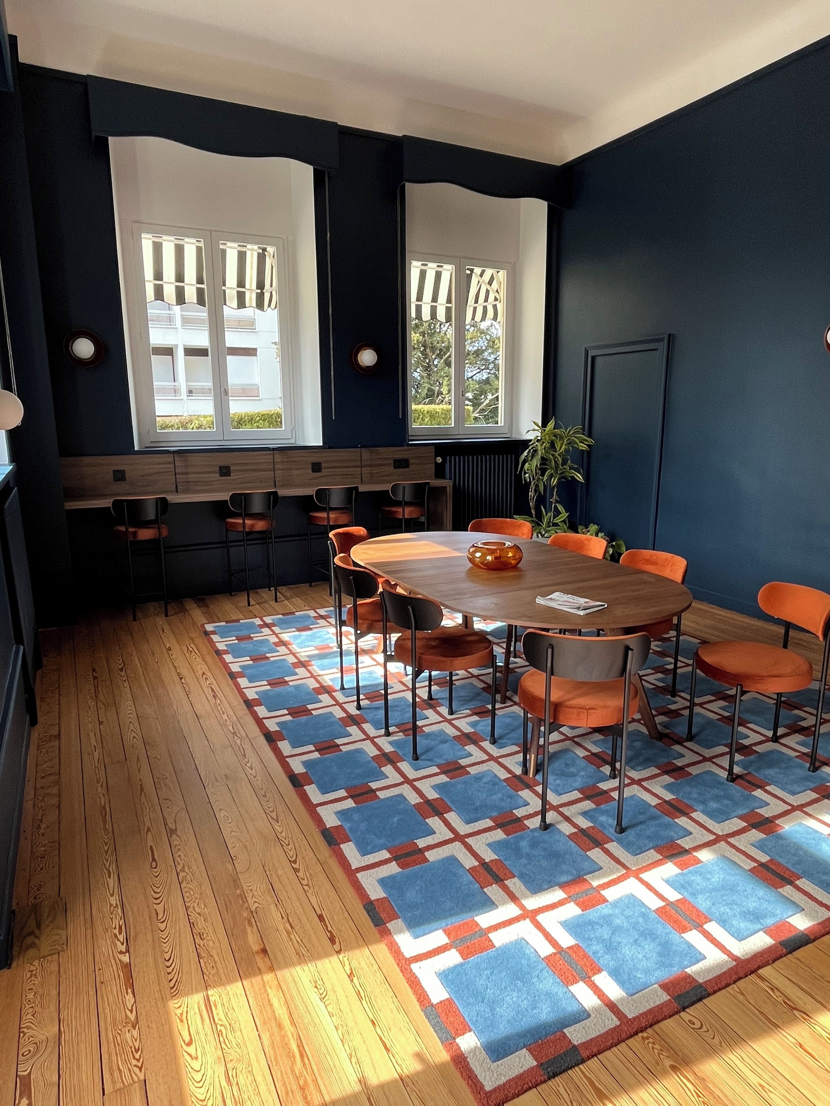 Un salón de conferencias con una mesa redonda, sillas naranjas, una alfombra estampada y ventanas amplias con cortinas.