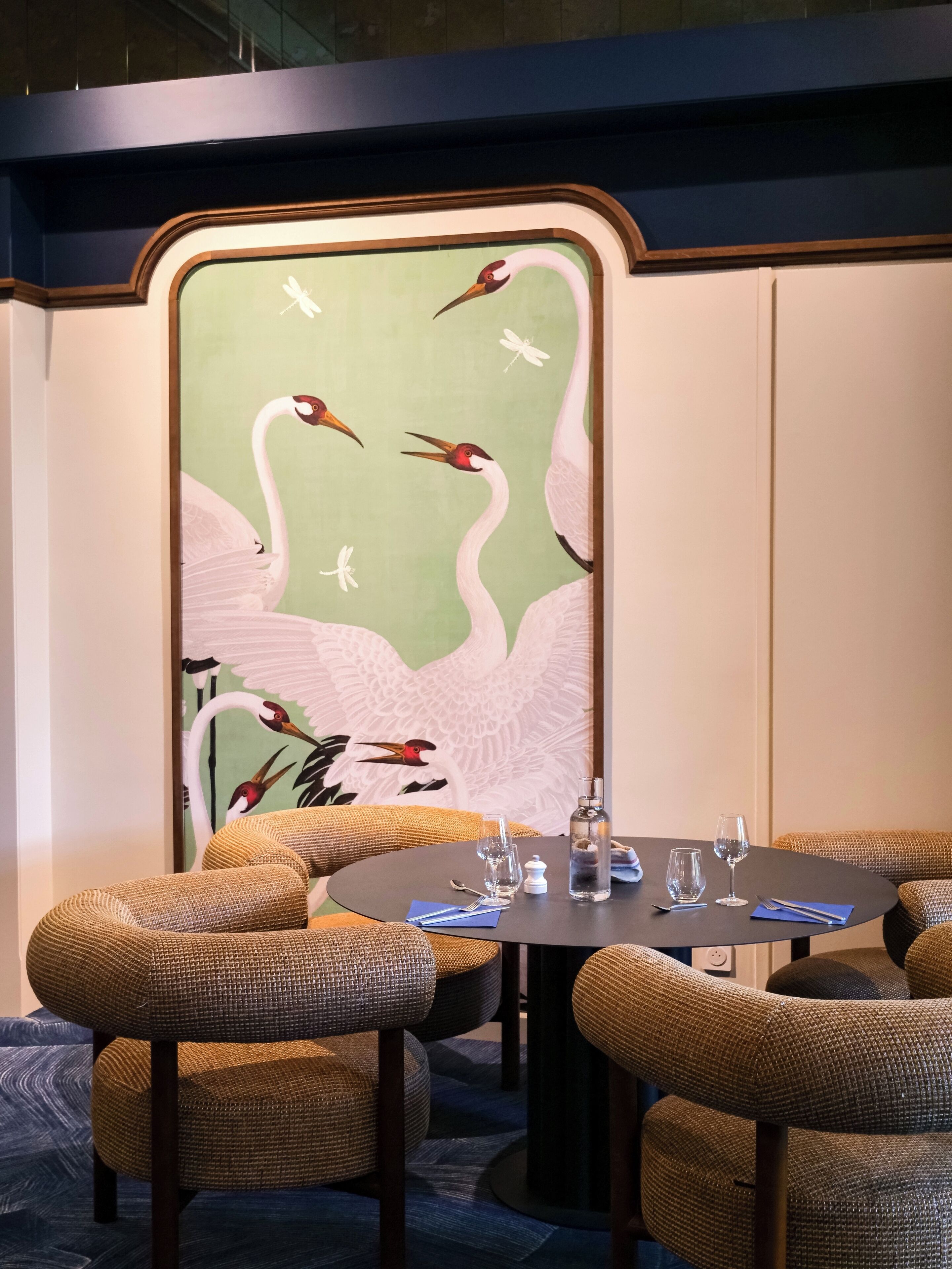 Un comedor con una mesa redonda dispuesta para cuatro personas, rodeada de sillas de mimbre. Un gran mural en la pared muestra pájaros blancos y una libélula.
