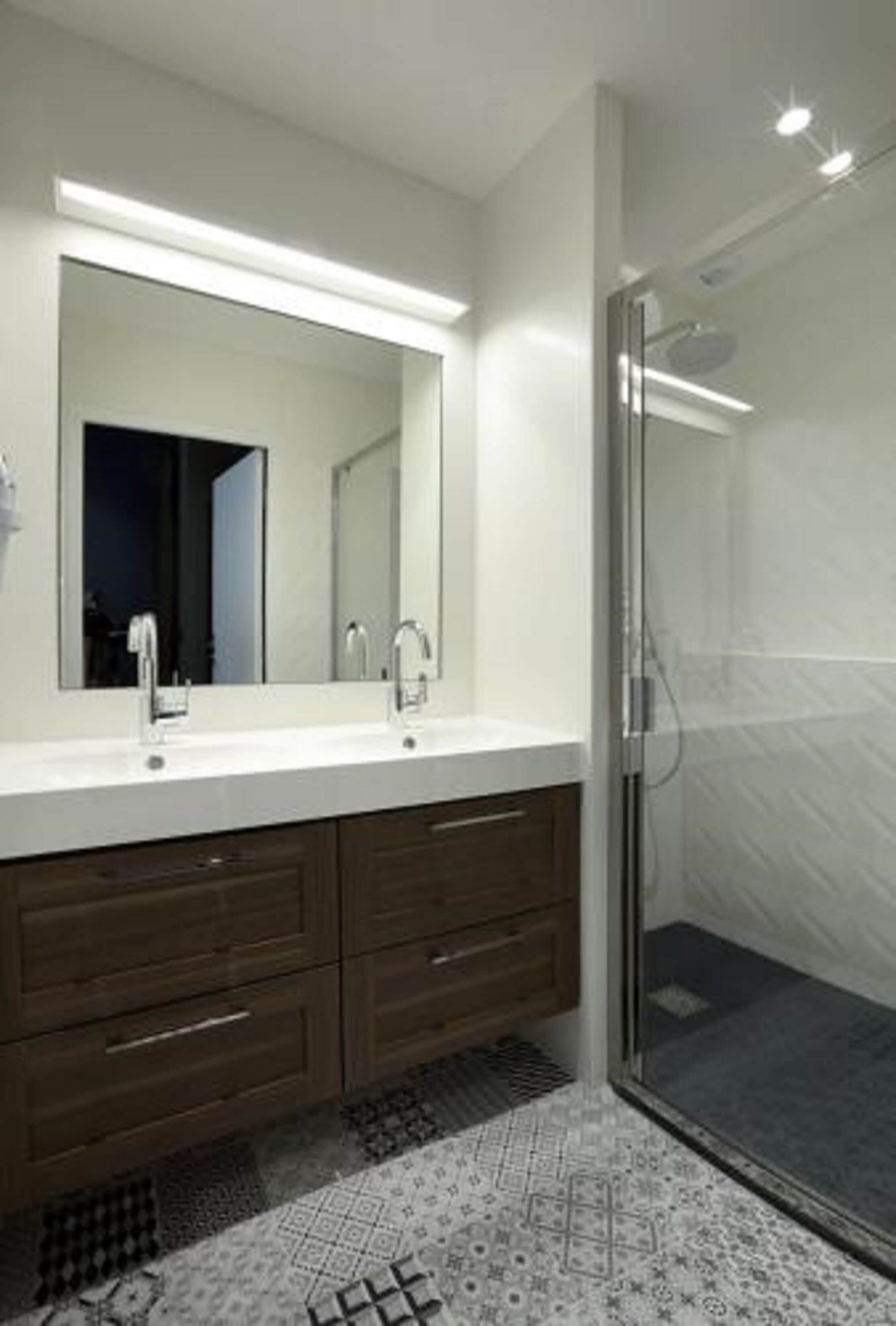 A modern bathroom with a large mirror, two sinks, a shower, and dark wooden cabinets.
