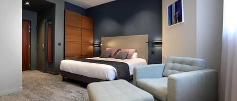 A modern bedroom with a large bed, two armchairs, a small ottoman, and a painting on the wall.