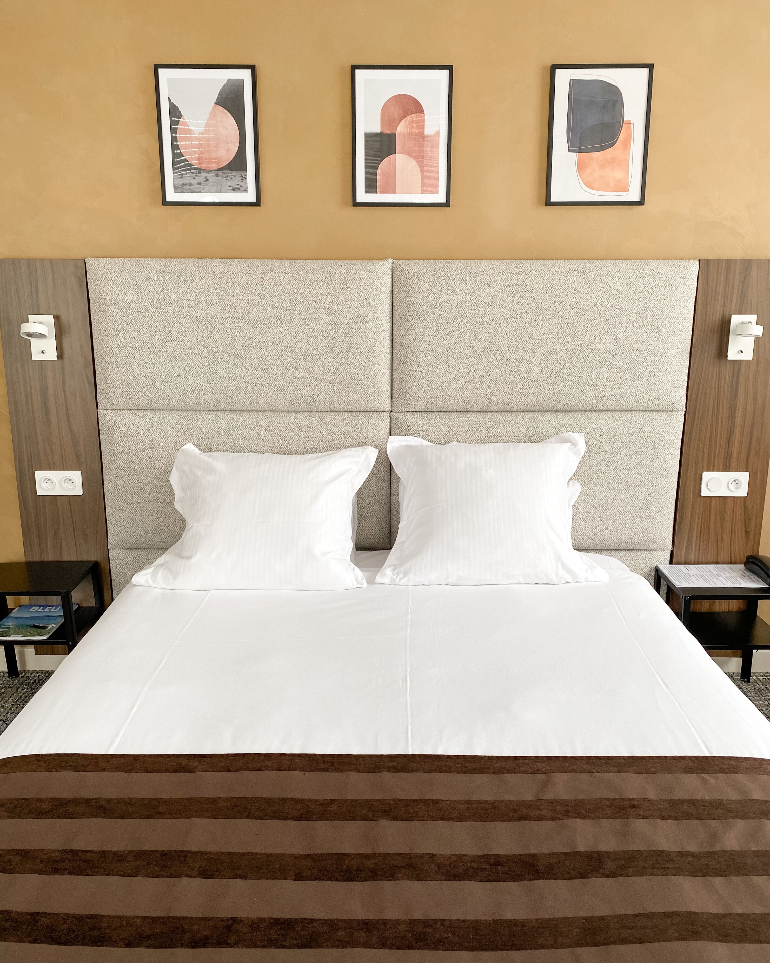Une chambre d’hôtel avec un grand lit, deux oreillers blancs, une tête de lit à motifs et trois œuvres d’art encadrées accrochées au mur.