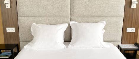 A hotel room with a large bed, two white pillows, a headboard with a textured pattern, and three framed artworks on the wall.