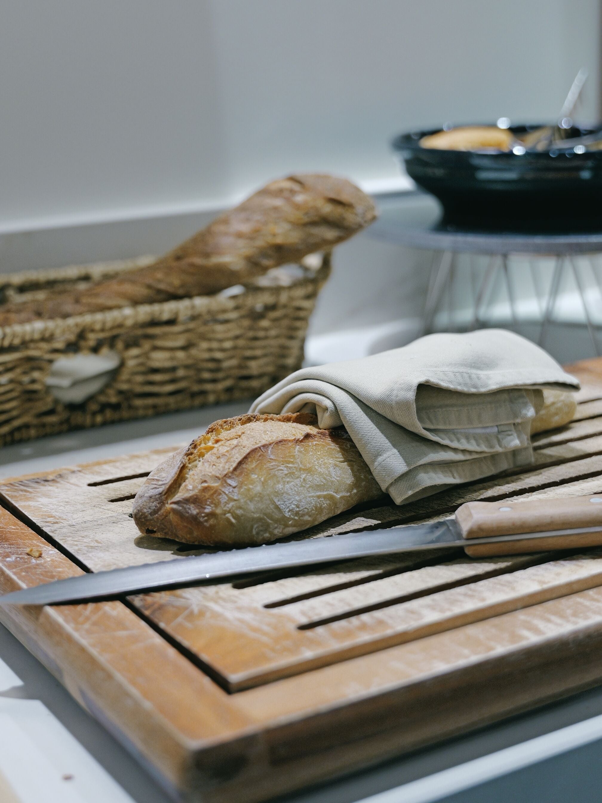 A loaf of bread on a wooden cutting board with a knife, a cloth, and a basket in the background.