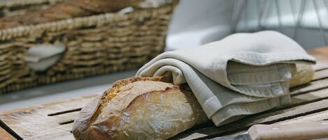 A loaf of bread on a wooden cutting board with a knife, a cloth, and a basket in the background.