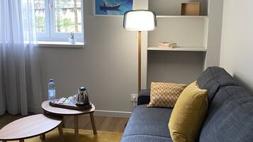 A living room with a grey sofa, a small round table, a lamp, a bookshelf, and a poster of a river scene.