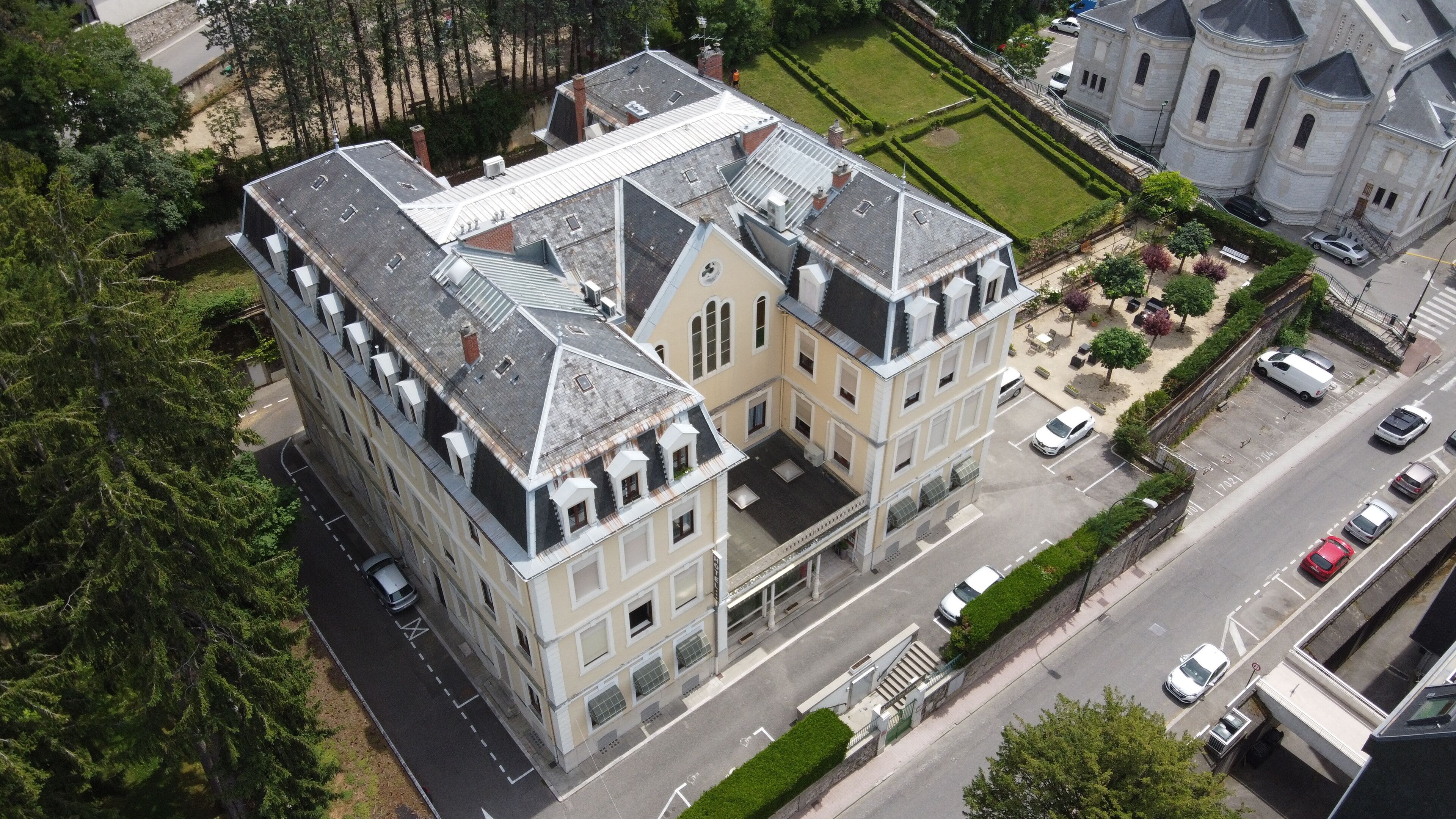 Vue aérienne d’un grand bâtiment historique à plusieurs étages, doté d’une cour et d’un espace de stationnement.