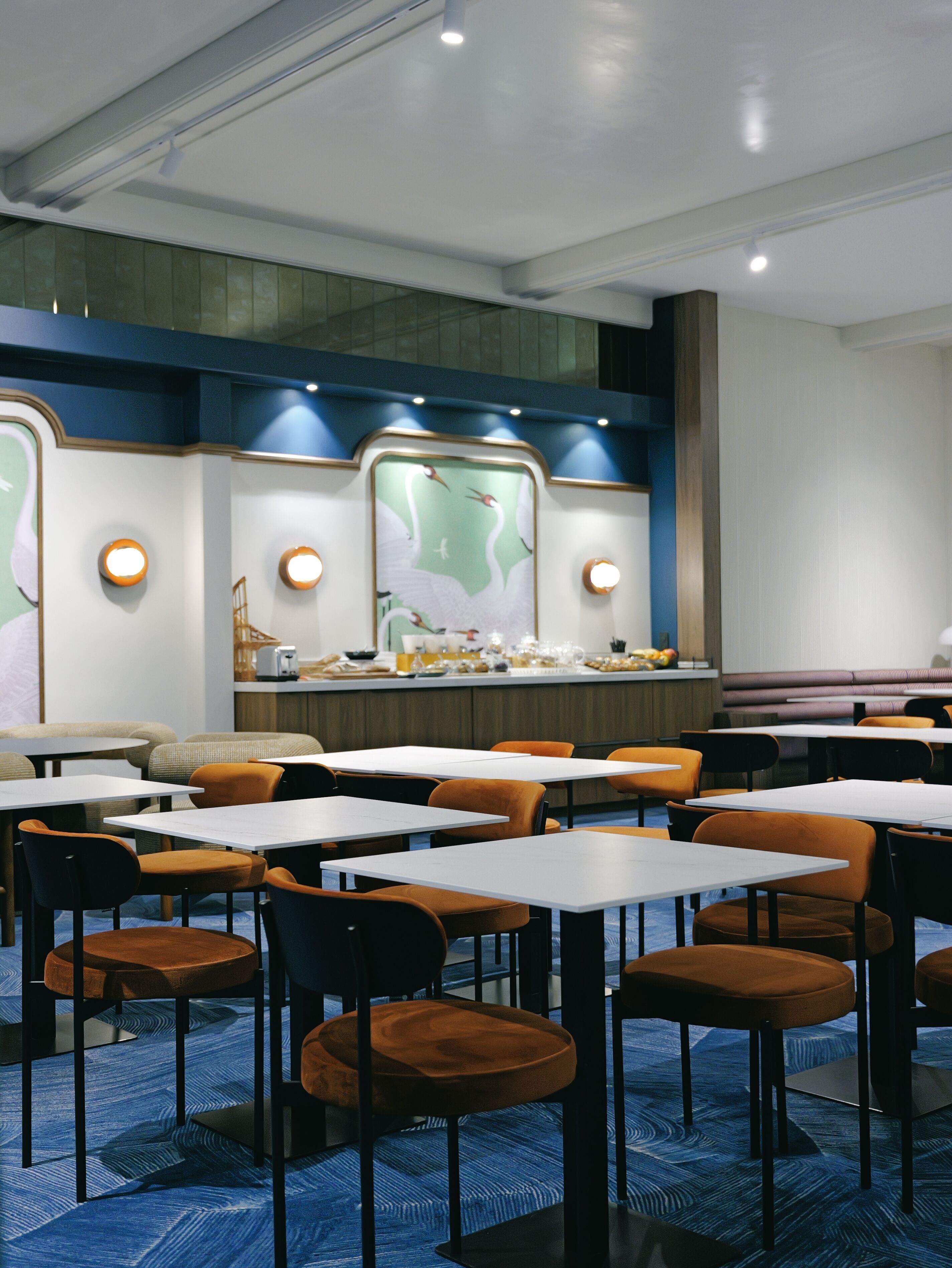 Un restaurant avec des tables et des chaises, un comptoir où sont exposés des plats, et un mur orné d’une fresque.