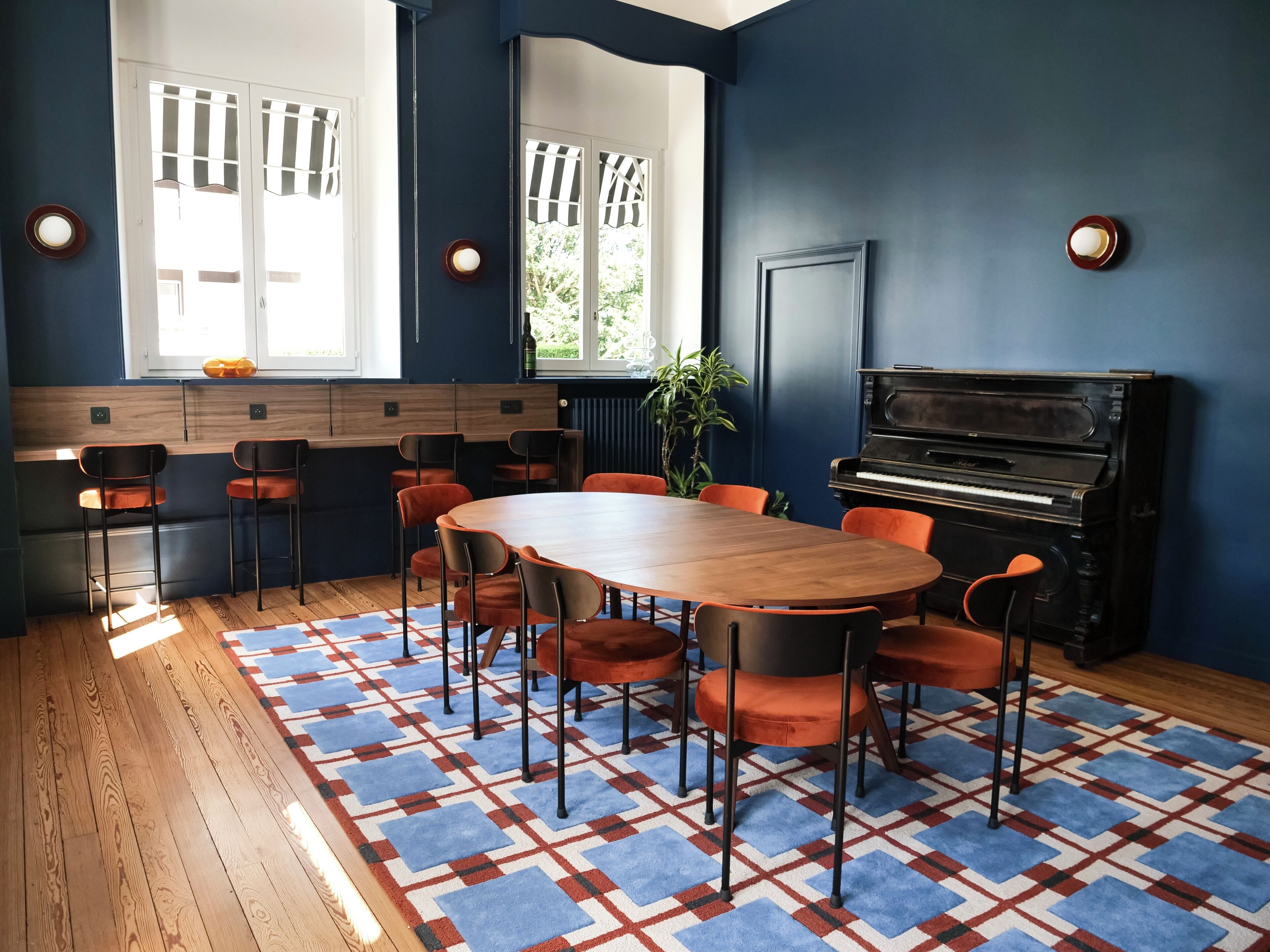 A room with a wooden table, orange chairs, a piano, and a patterned rug.